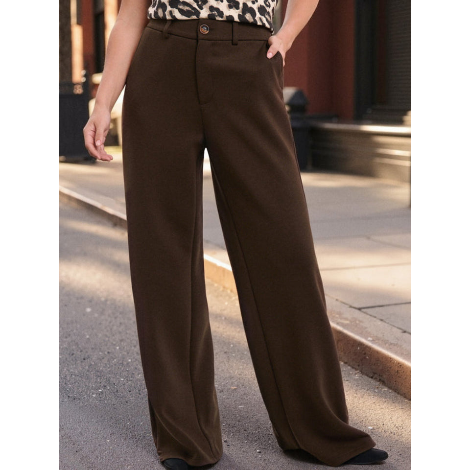 A person wearing dark brown wide-leg trousers, standing outdoors on a sidewalk with one hand in a leopard-print top.