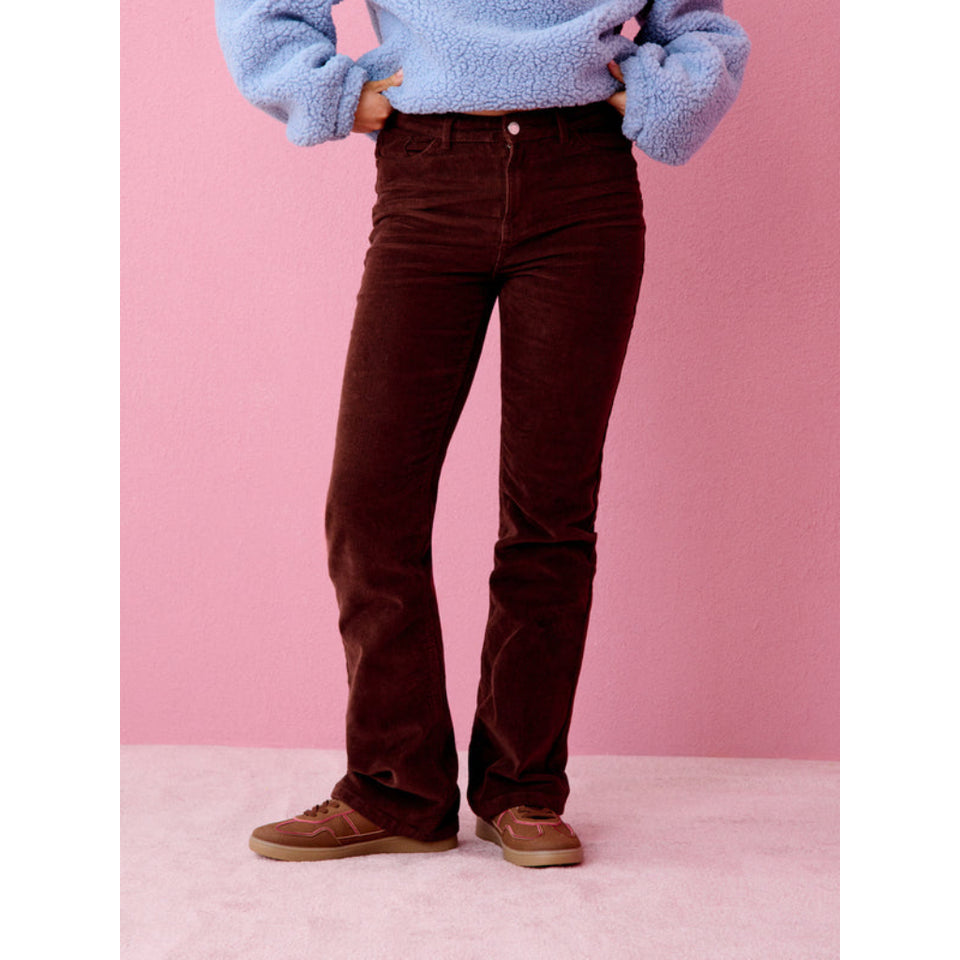 A person wearing dark brown corduroy flare jeans, a light blue fuzzy sweater, and brown sneakers, standing against a pink background.