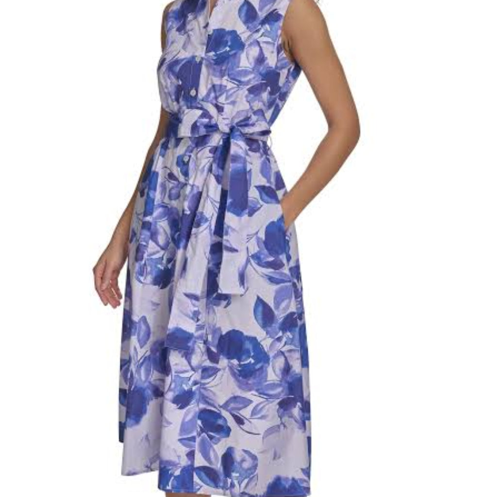 A woman modeling a sleeveless midi dress with a blue and white floral print, featuring a wrap-style bodice and a self-tie belt, paired with white strappy heeled sandals against a white studio background.