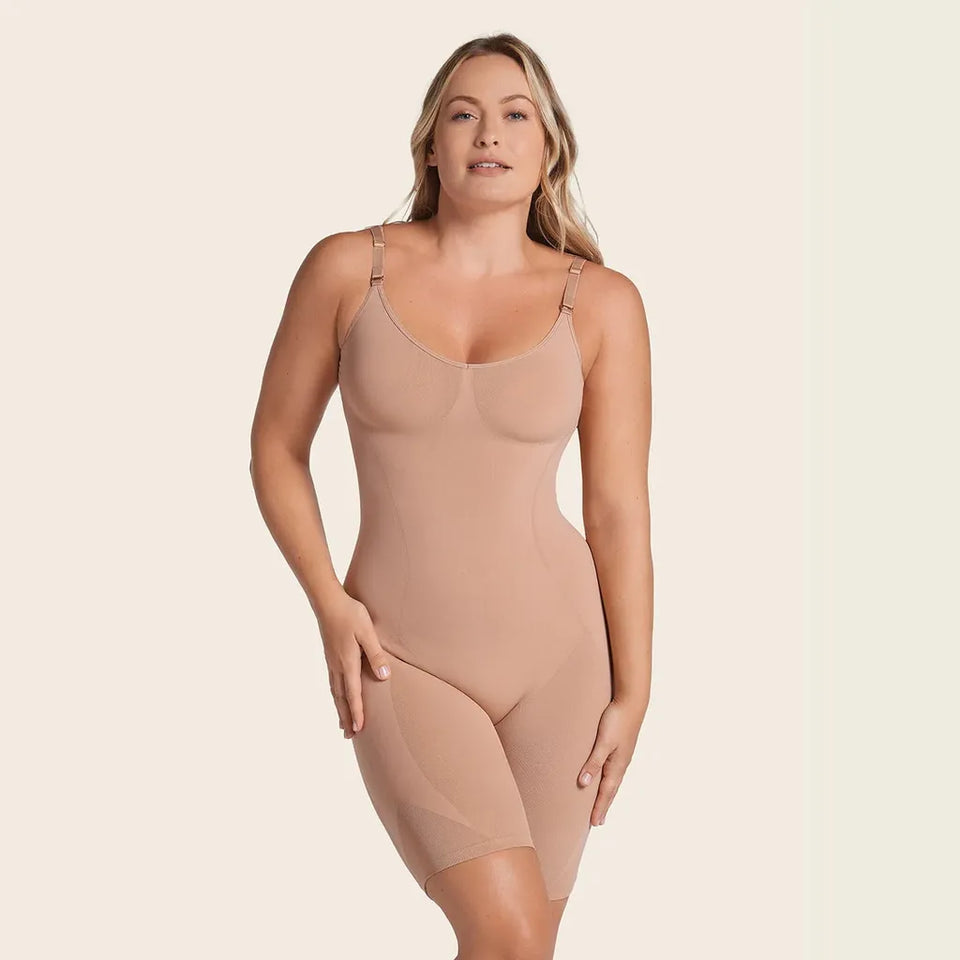 A woman modeling a nude-beige compression bodysuit with shoulder straps and mid-thigh legs against a solid light beige background.