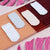 Four pairs of ankle socks in white, light blue, cream, and dark red are displayed on a geometric, slatted platform with pink decorative props in the background.