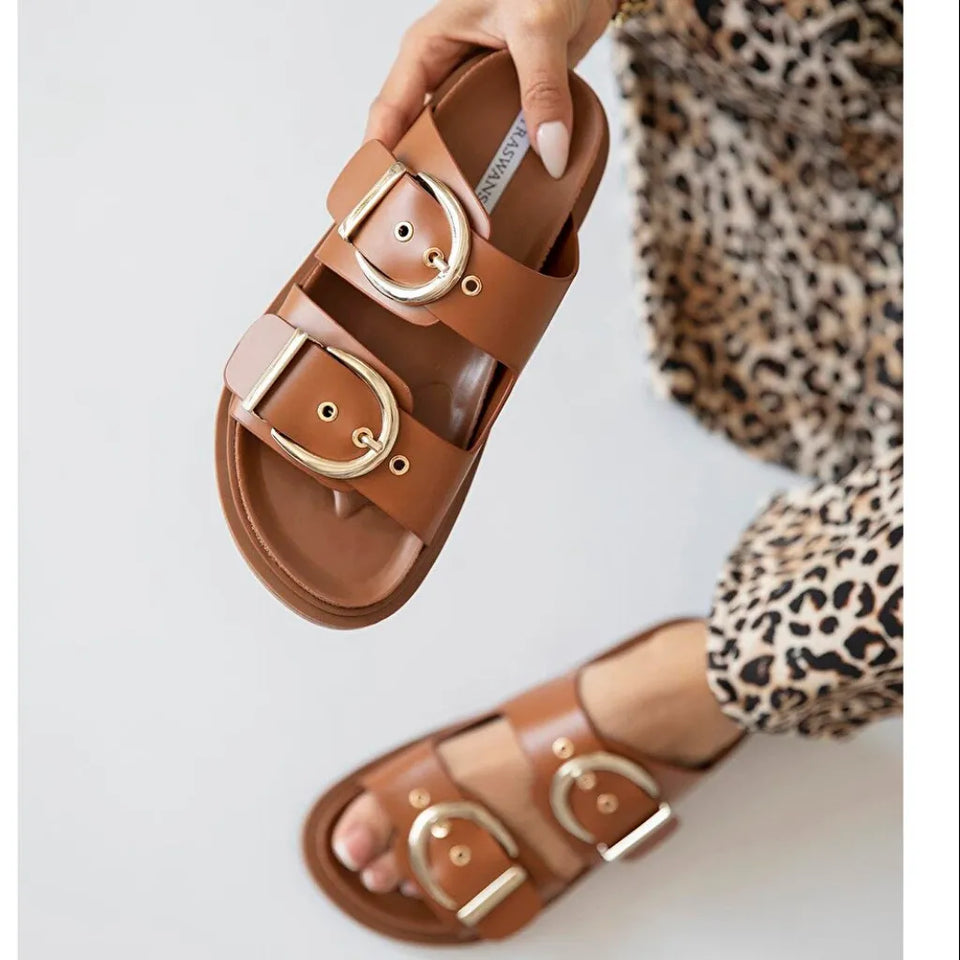 A person holds up a tan double-strap slide sandal with large gold-tone oval buckles against a light gray background, with the matching sandal also visible on the foot and a leopard-print garment shown.