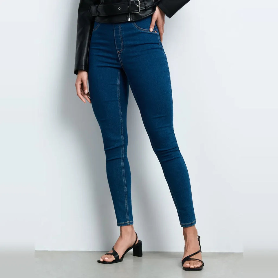 A model wearing dark blue skinny jeans, styled with a black leather belt, black leather jacket, and black strappy block-heel sandals, standing against a light gray studio background.