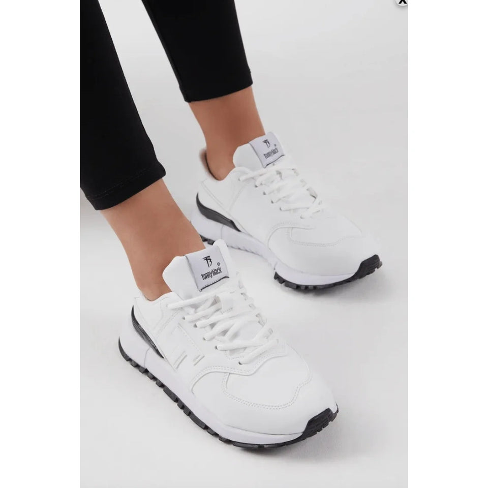 White low-top sneakers with black outsole accents, shown on a model wearing black leggings against a light gray studio background.