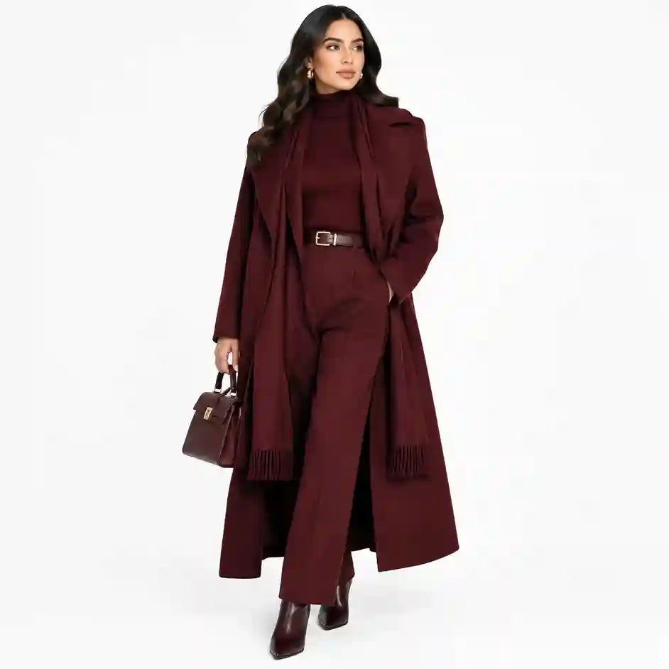 A woman models a matching three-piece winter outfit in deep burgundy, featuring a long coat with a scarf, a turtleneck sweater, and wide-leg trousers, styled with a brown belt and holding a matching handbag against a white background.