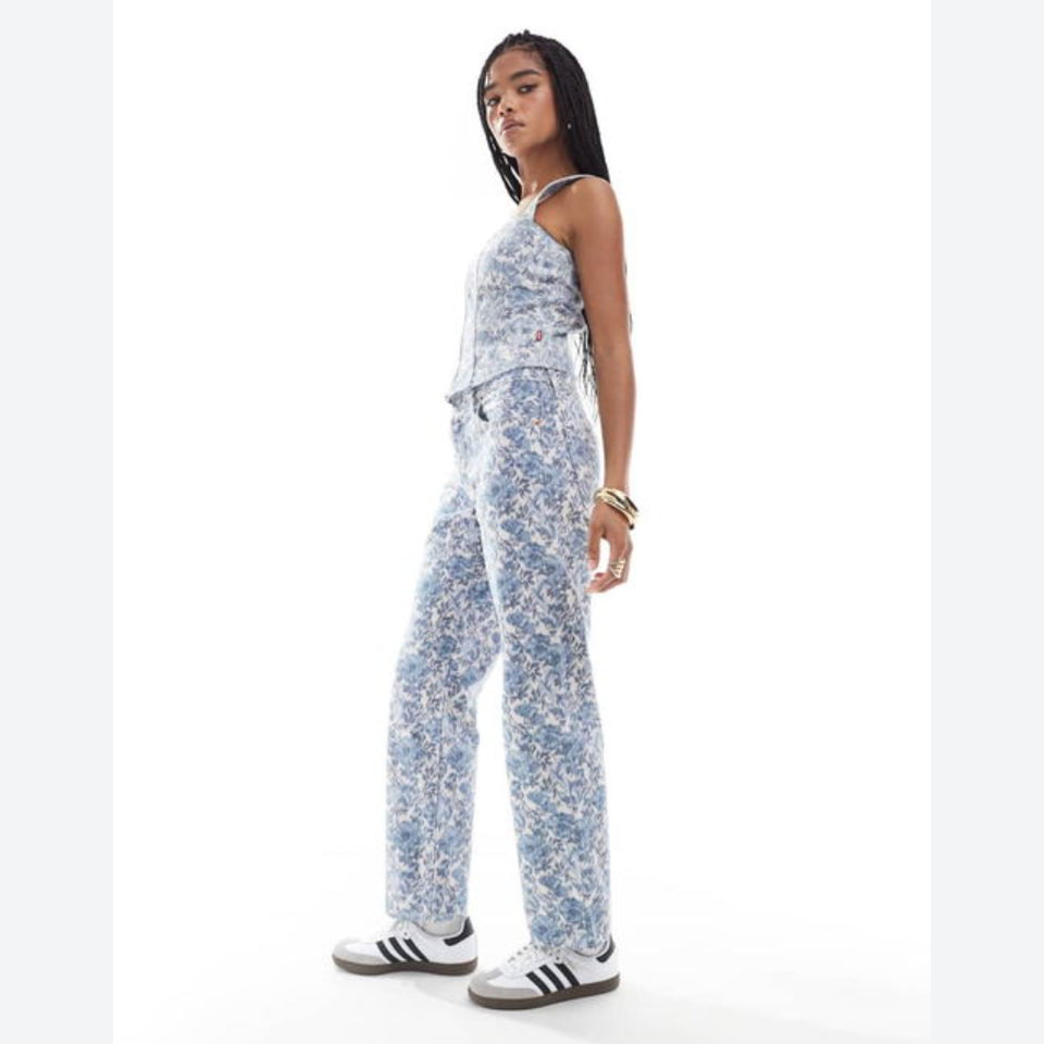 A woman modeling a sleeveless light blue and white floral print jumpsuit, paired with white sneakers, against a plain white studio background.