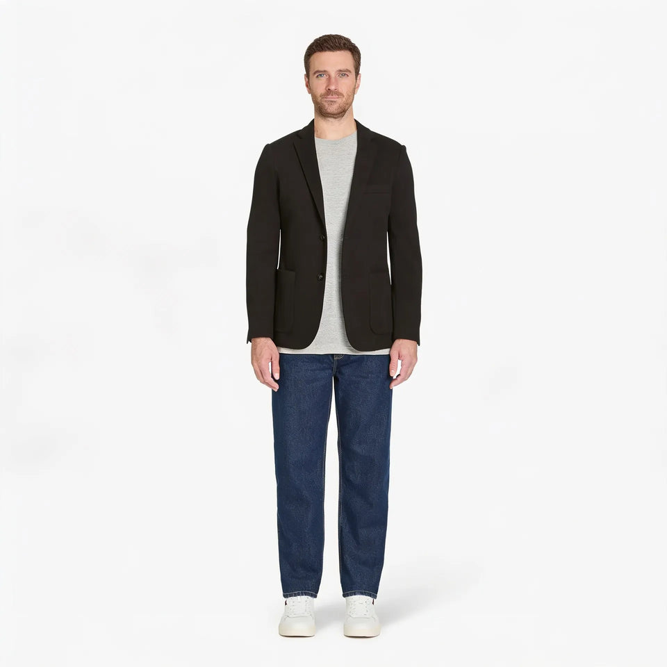 Man wearing a black blazer, gray shirt, blue jeans, and white sneakers on a white background