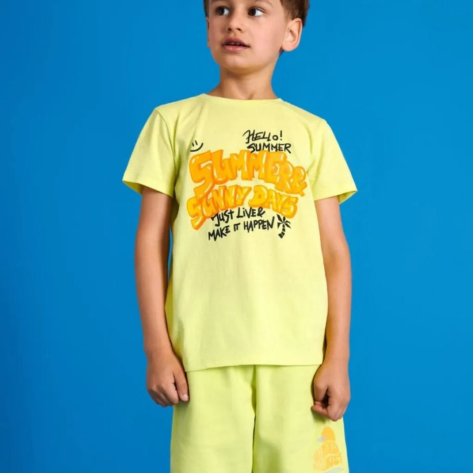 A young boy wearing a bright yellow-green t-shirt and matching shorts set, featuring a colorful graphic on the front that reads 'HELLO! SUMMER STUDY DAYS', posed against a solid blue background.