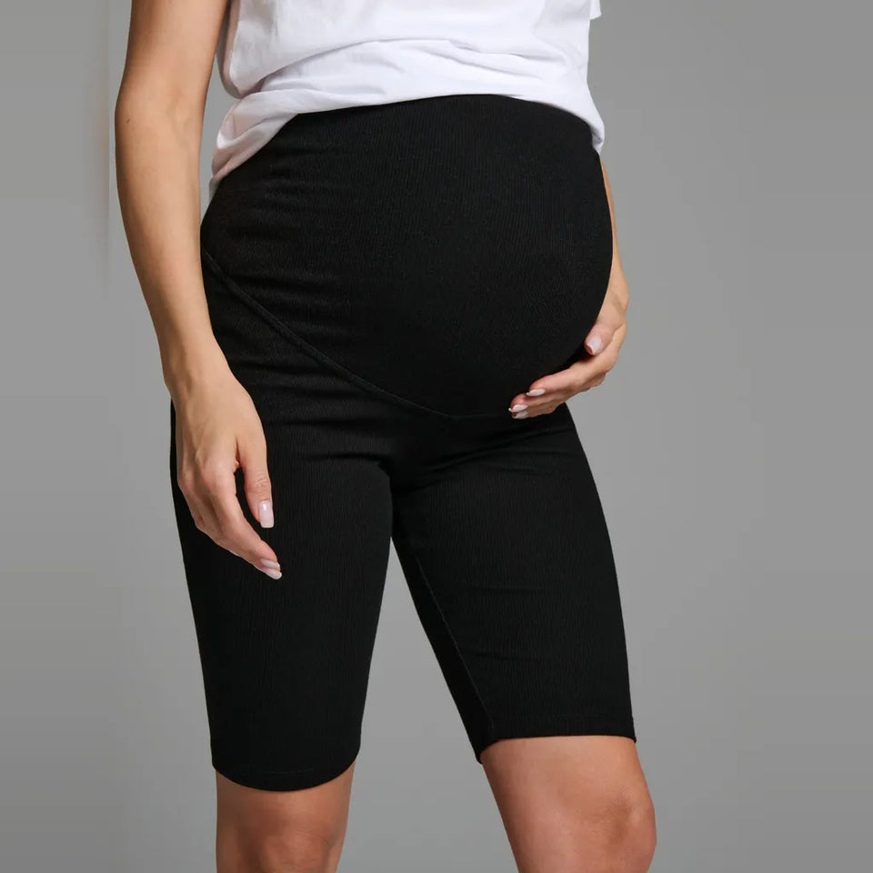 A pregnant model wearing black maternity biker shorts with a built-in belly support pouch, paired with a white t-shirt, against a solid gray studio background.