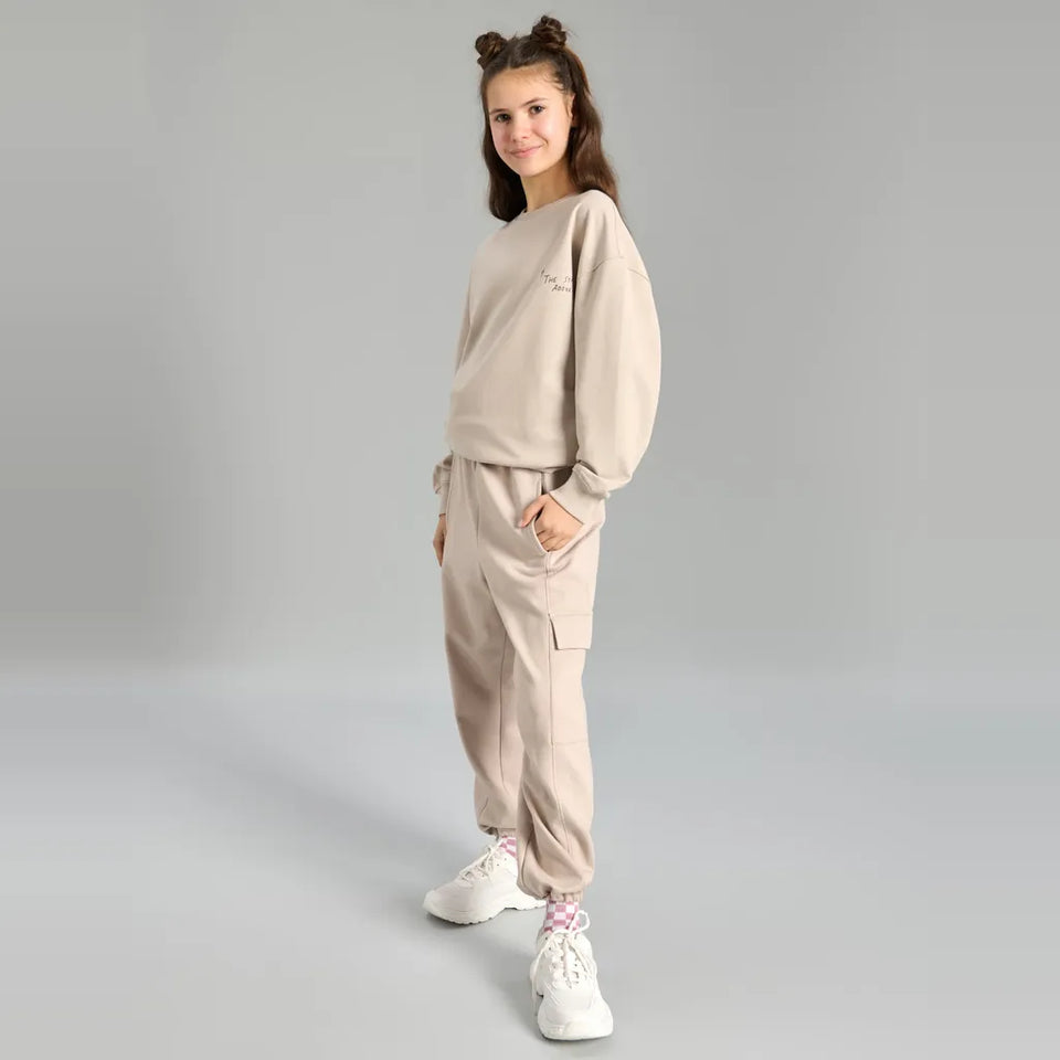 A young girl modeling a light beige two-piece lounge set consisting of a sweatshirt and matching jogger pants, paired with white sneakers, against a neutral gray studio background.