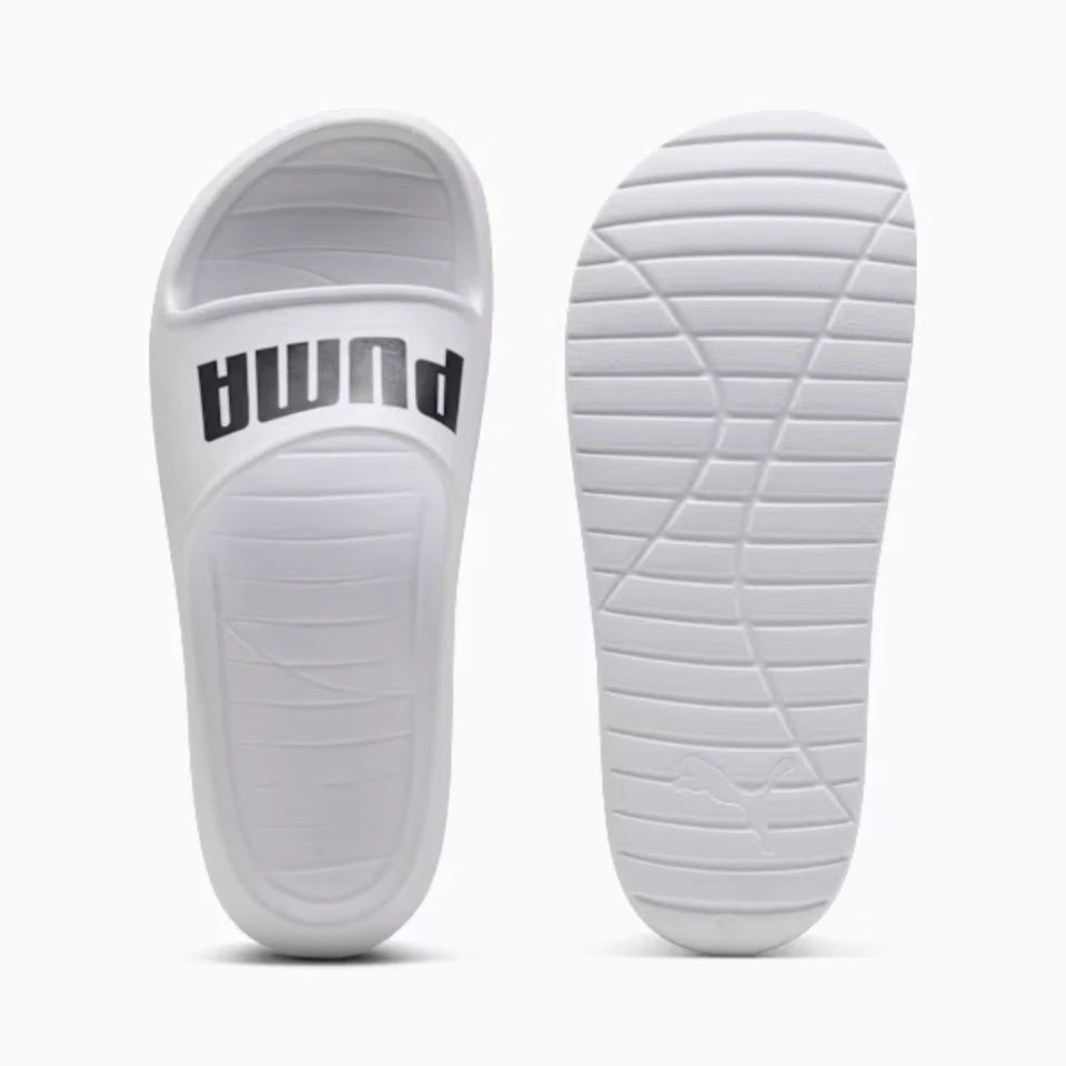 Two white Puma slide sandals shown on a light gray background, with one sandal displaying the side profile with the black Puma logo and the other showing the treaded outsole.