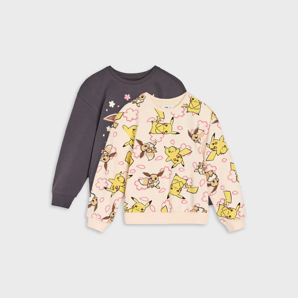 Two children's long-sleeve crewneck sweatshirts laid flat on a light gray background. The front sweatshirt is light pink with a repeating print of yellow Pikachu and brown Eeveo characters outlined in pink.