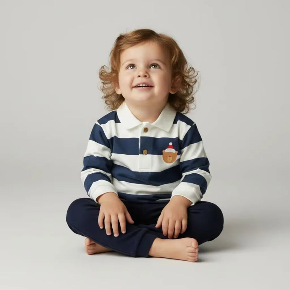 A toddler with curly light brown hair sits cross-legged on a light gray studio background, wearing a navy and white horizontally striped long-sleeve polo shirt with a small cupcake graphic on the chest, paired with navy pants.