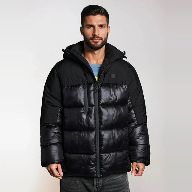 A man wearing a black hooded puffer jacket with horizontal quilting, shown from the waist up against a light gray studio background.