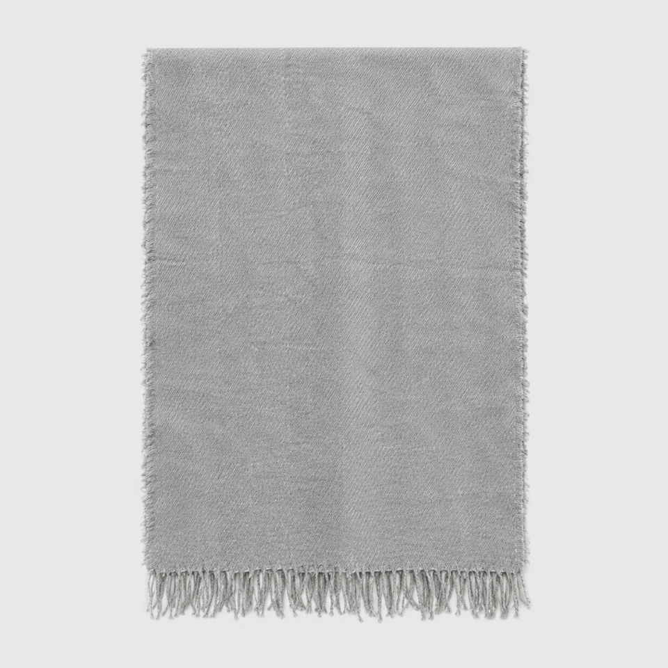A light gray woven throw blanket with fringed edges, displayed flat against a neutral light gray background.
