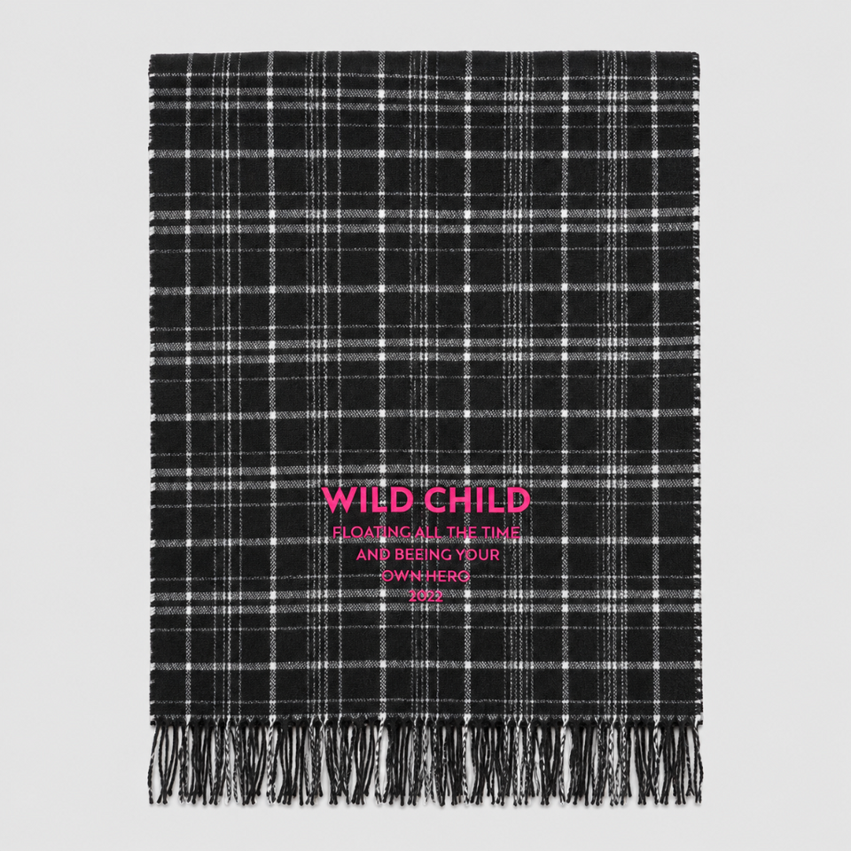A black and white plaid fringed throw blanket with pink text reading 'WILD CHILD' and 'SLOWING ALL THE TIME AND BEING YOUR OWN HERO 2022' on a light gray background.