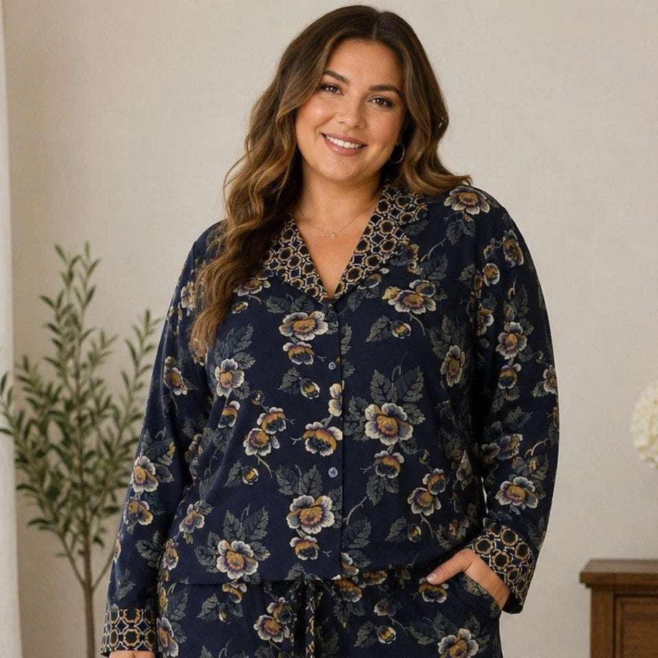 A woman modeling a two-piece navy blue pajama set with a floral print and patterned trim, standing in a cozy bedroom setting.