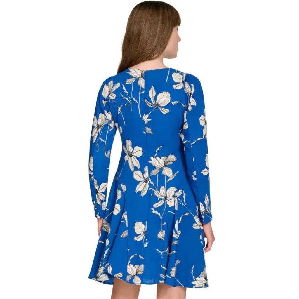 A woman shown from the back wearing a long-sleeve blue dress with a white floral print, set against a plain white background.