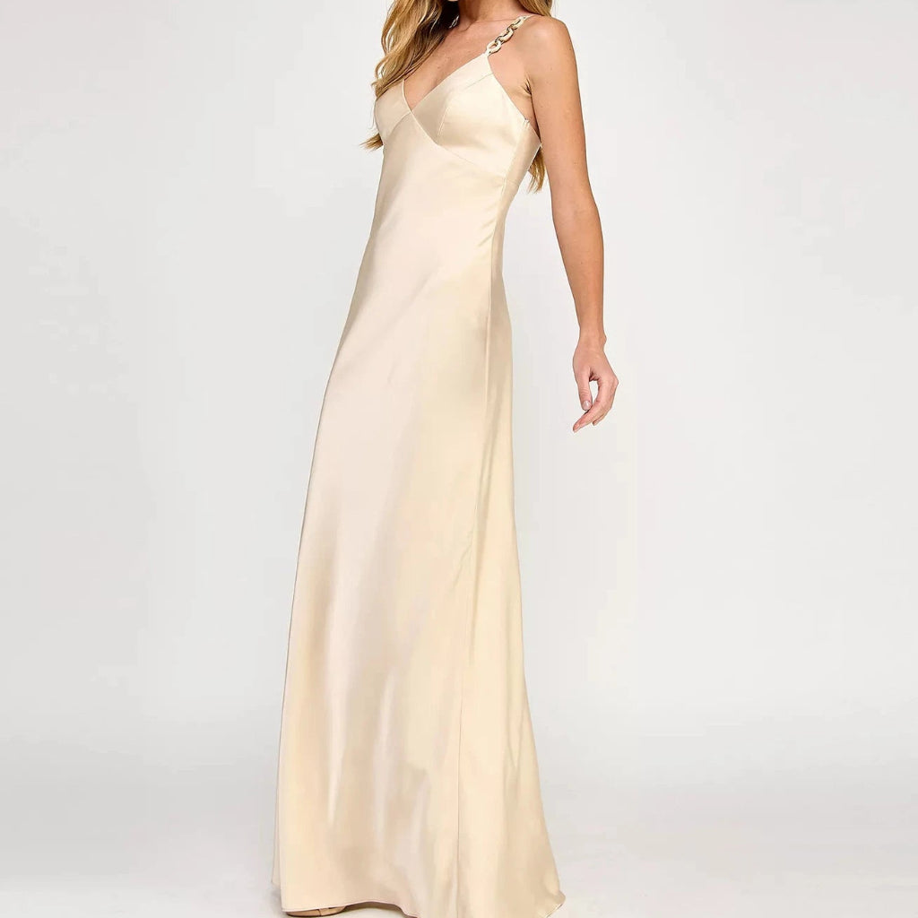A woman modeling a floor-length champagne satin gown with a V-neckline and a single strap accented with a crystal embellishment, set against a plain light gray background.