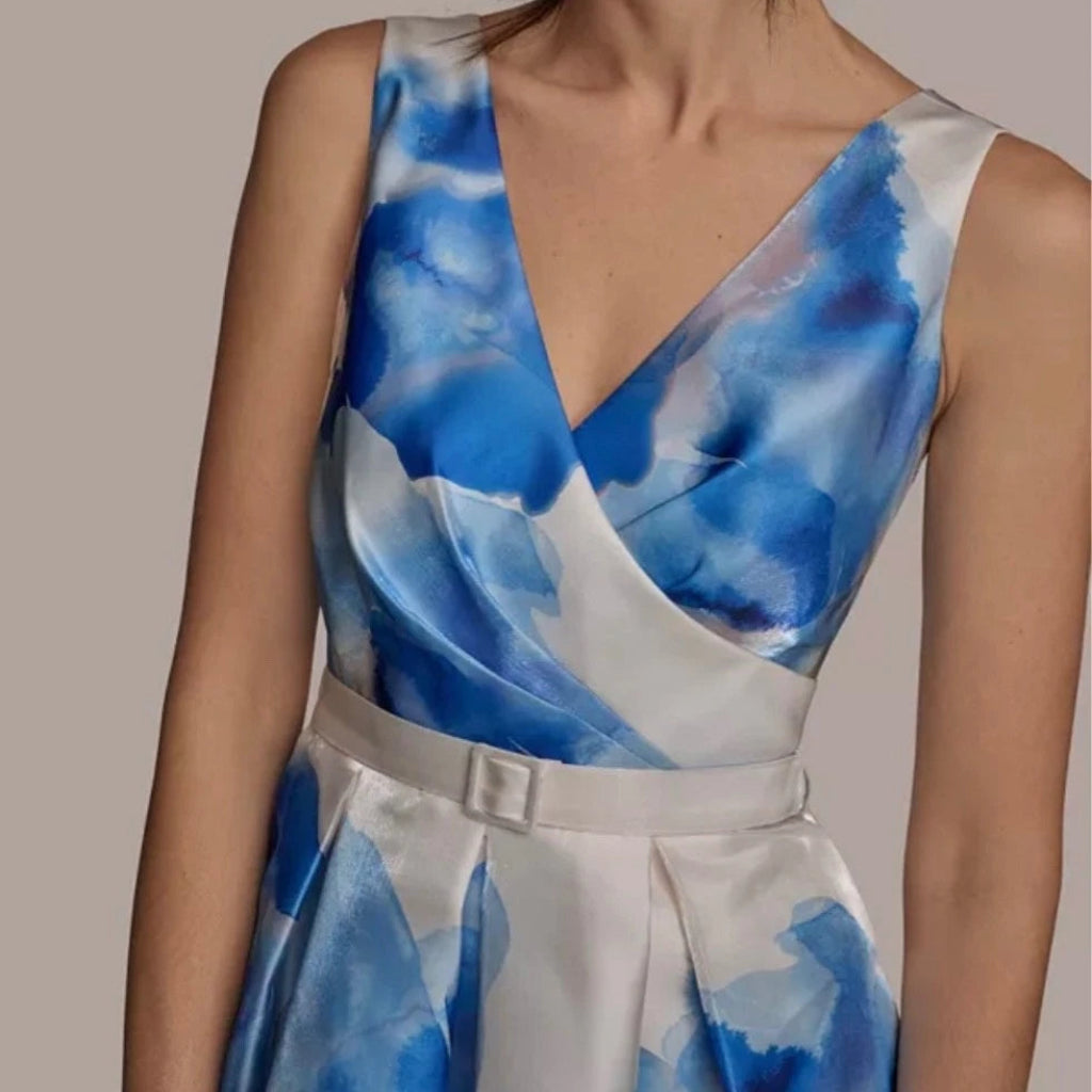 A woman wearing a sleeveless wrap-style dress with a blue and white watercolor print and a matching waist belt, shown against a plain taupe background.