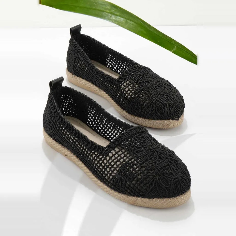A pair of black crocheted slip-on espadrille-style shoes with natural jute-wrapped soles, photographed on a clean white surface with a green leaf prop in the background.