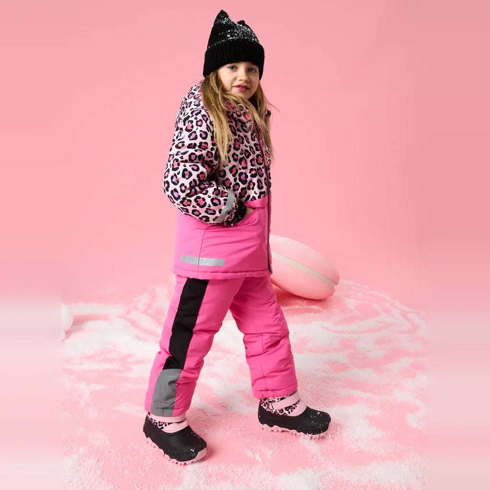 A young girl wearing a pink and black snowsuit, a leopard-print puffer jacket, a black knit beanie, and black snow boots, standing in a pink studio setting with a pink ball prop.