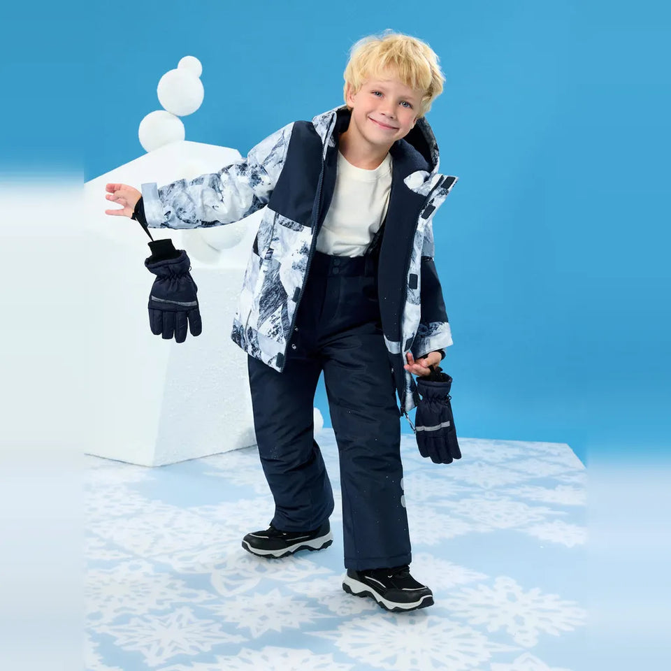 A young blonde boy modeling a winter ski outfit, including a patterned jacket, dark pants, gloves, and sneakers, standing in front of a blue studio background with a snowman prop.