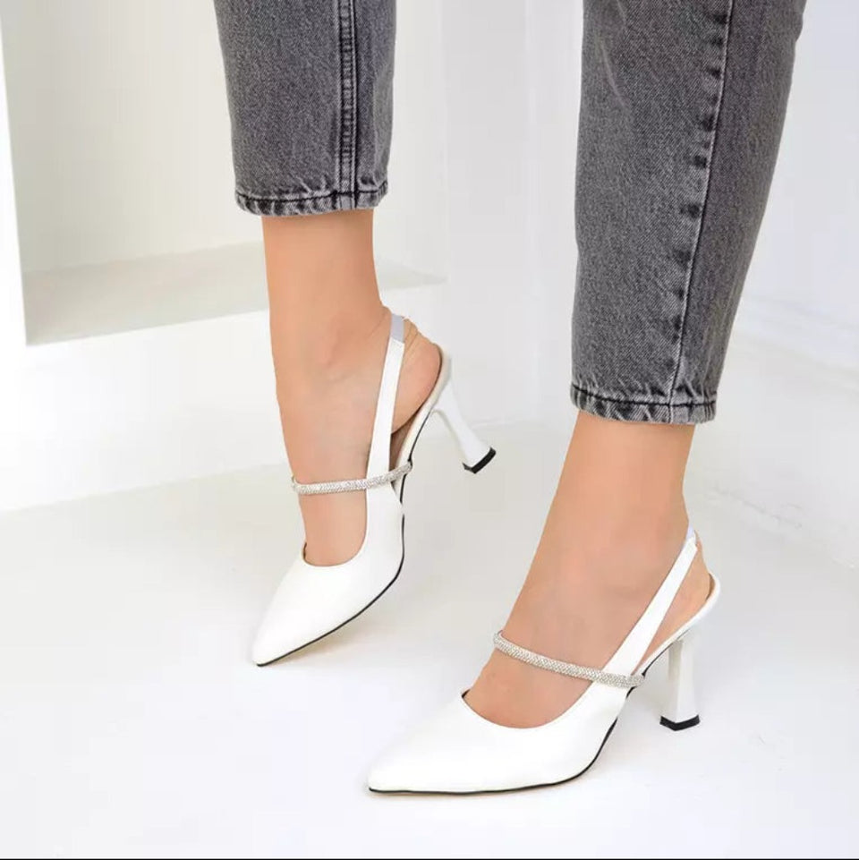 White slingback pointed-toe high heels with rhinestone-embellished straps, shown on a model wearing gray cuffed jeans against a clean white background.