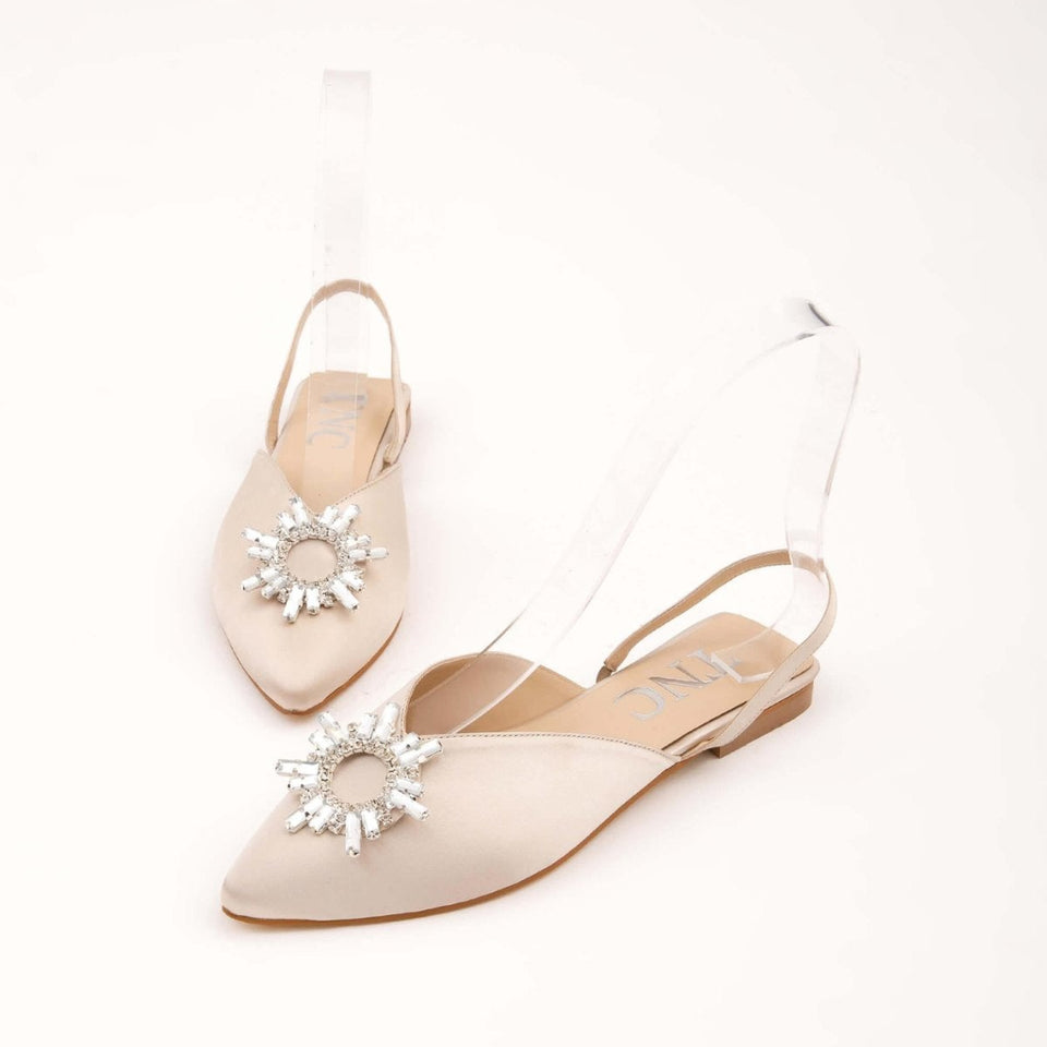 A pair of pointed-toe slingback flats in a light nude beige satin, embellished with a clear rhinestone sunburst flower on the toe and a transparent strap, shown on a clean off-white background.
