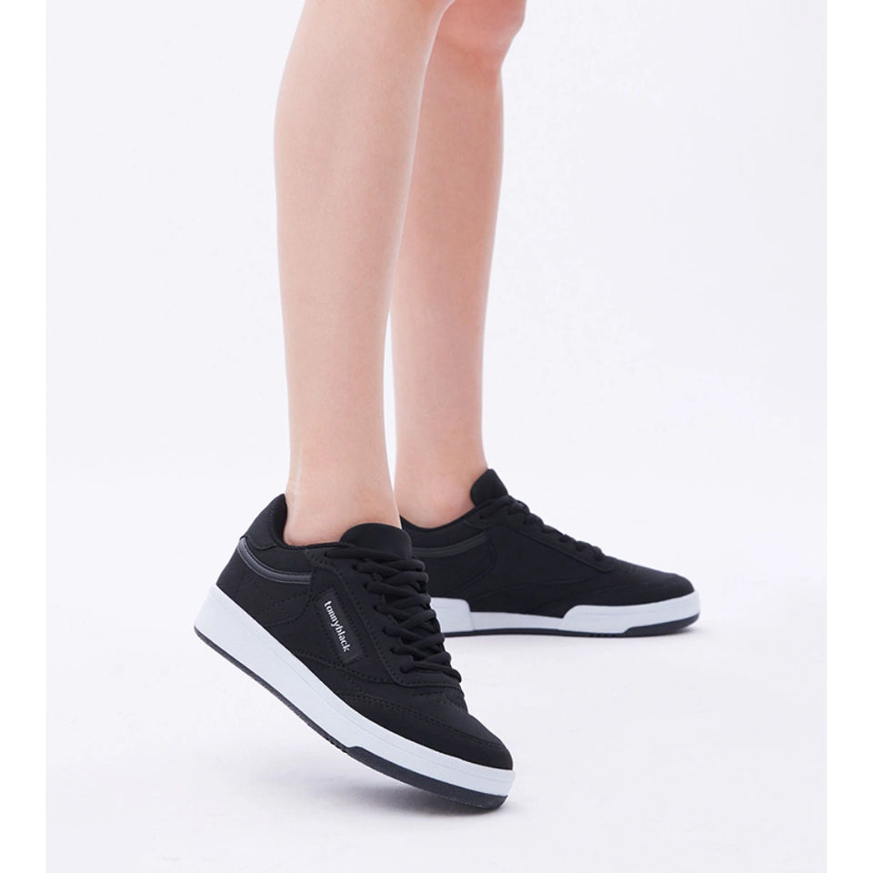 A pair of black low-top sneakers with white midsoles, shown on a model's feet against a clean white studio background.