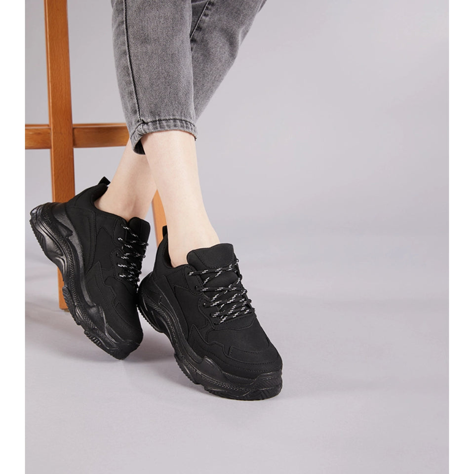 A person wearing black chunky lace-up sneakers with gray cuffed jeans, standing on a light gray floor next to a wooden stool.