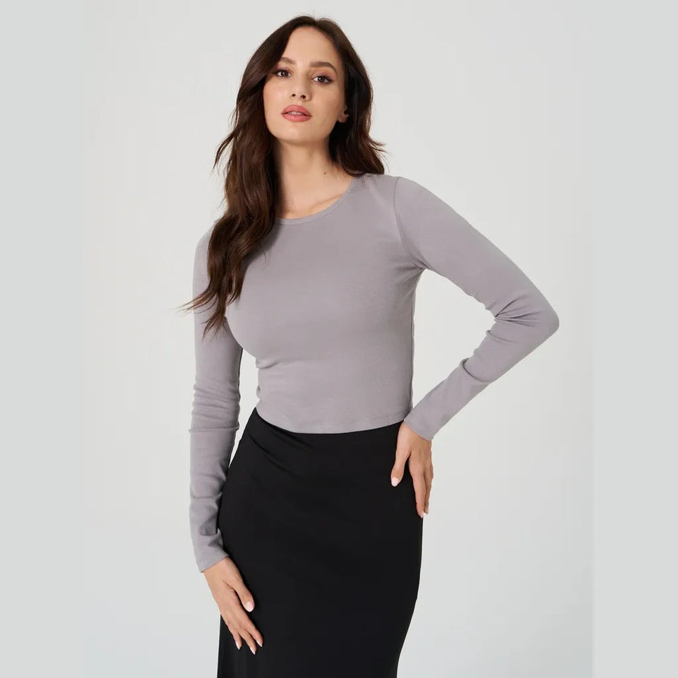 A woman modeling a fitted, long-sleeve crewneck top in a light gray color, paired with a black skirt, against a plain light gray studio background.