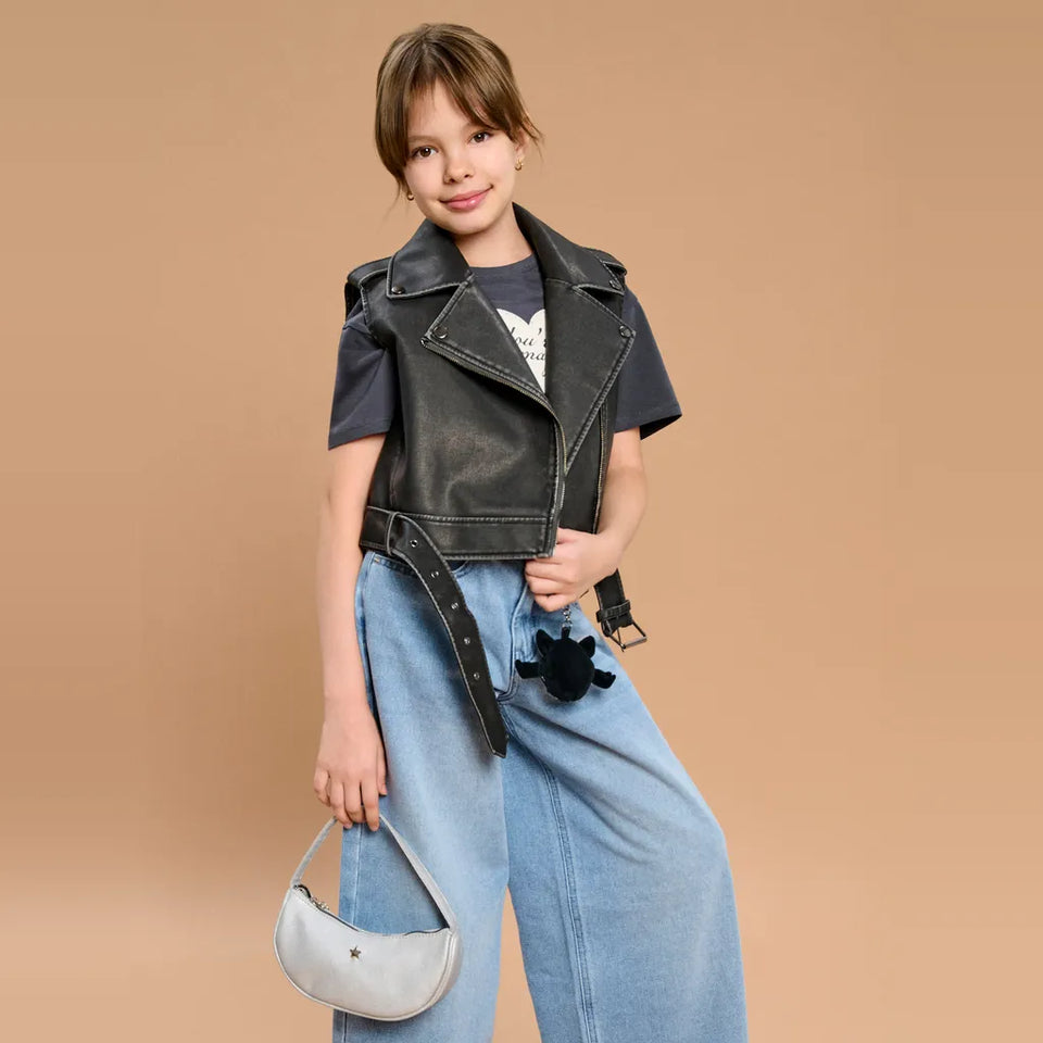 A young girl modeling a black faux-leather moto jacket vest over a dark t-shirt, paired with wide-leg light blue jeans, a black studded belt, and holding a small white crescent moon handbag against a solid tan background.