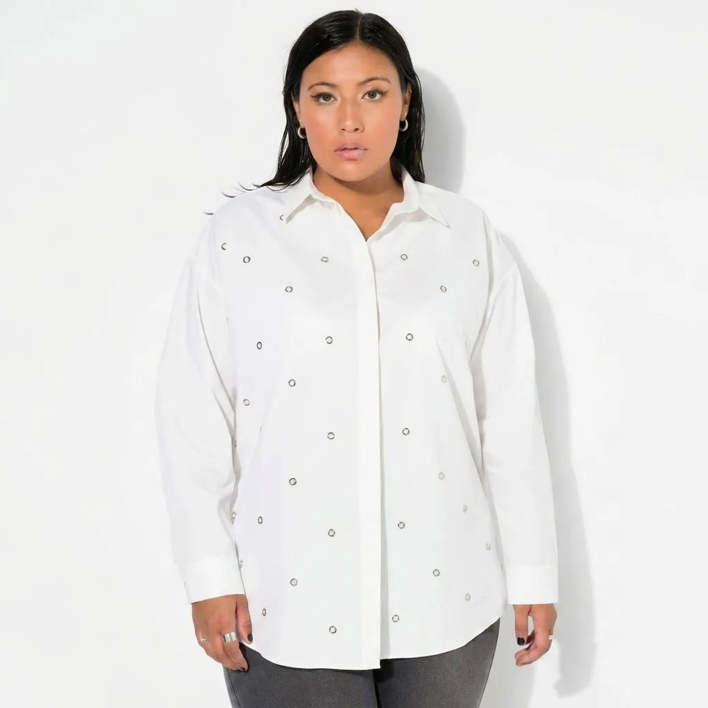 A woman modeling a white long-sleeve button-up shirt with smal silver stud embellishmentsl , paired with dark gray pants, against a plain white studio background.
