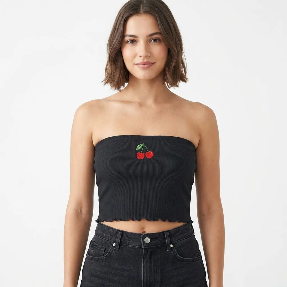 A woman modeling a black strapless ribbed crop top with a small red cherry and green leaf graphic on the chest, paired with dark denim jeans against a plain white background.