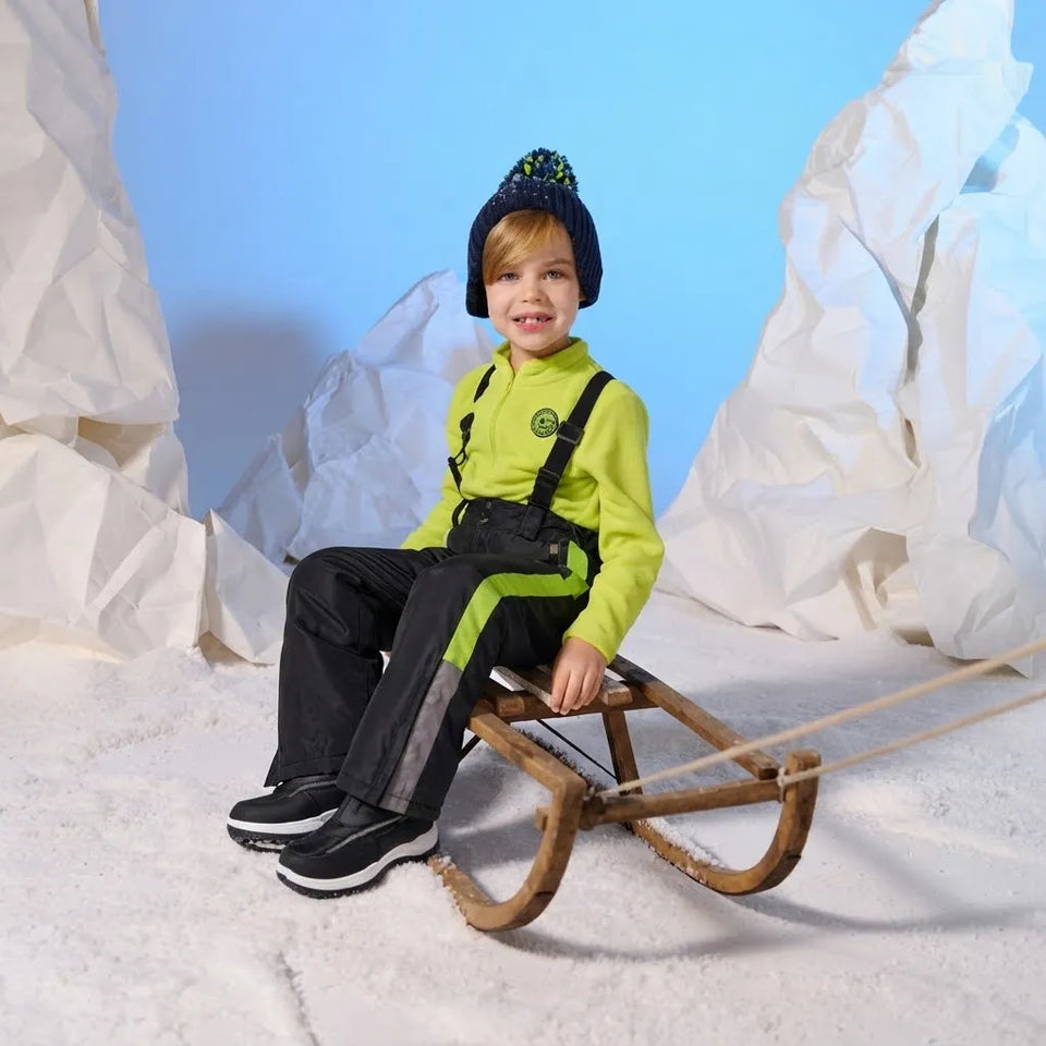 A young child wearing a bright lime green zip-up jacket, black snow pants with reflective stripes, and a dark knit beanie sits on a wooden sled in a studio set designed to look like snow with white paper mountains and a blue backdrop.