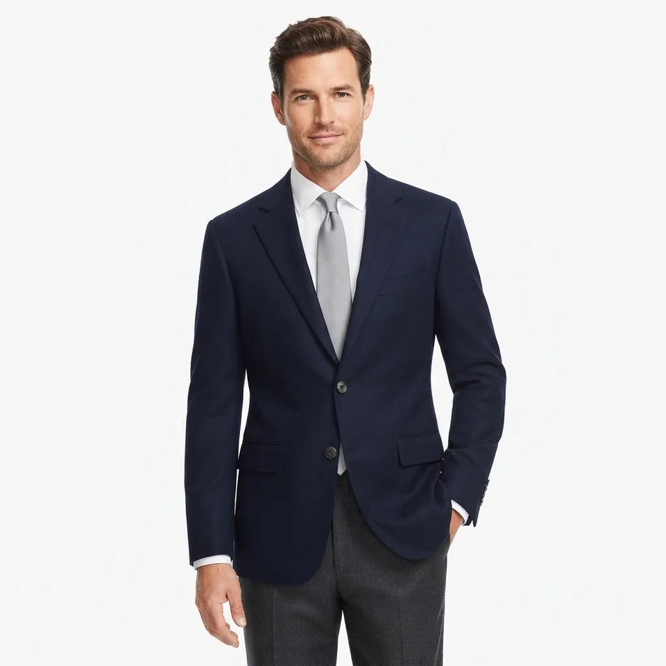 A man wearing a navy blue two-button suit jacket, a white dress shirt, a light gray tie, and gray trousers, standing against a plain white studio background.
